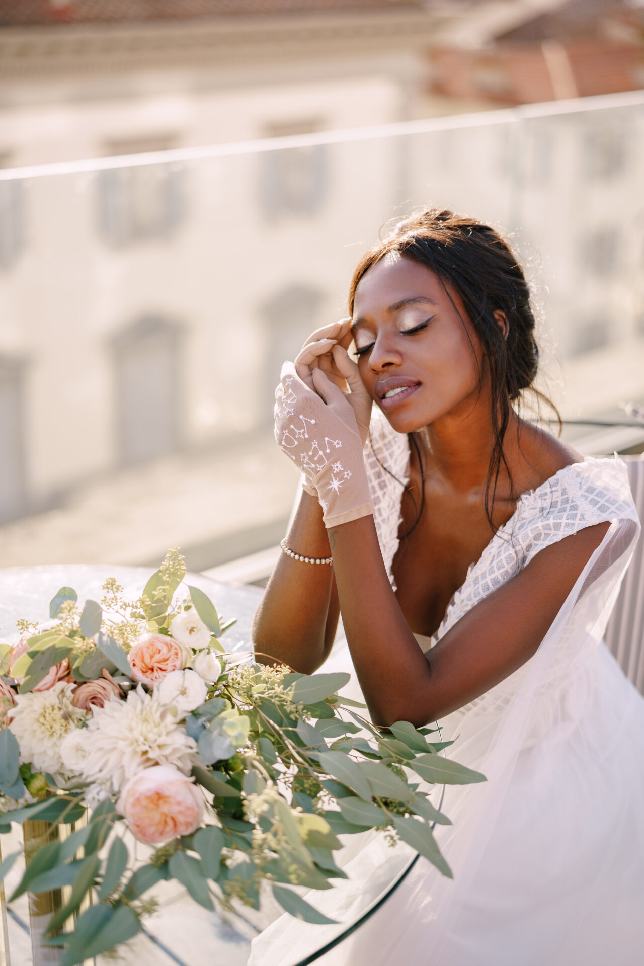 African-American bride sits at the table, touches her face with gloves in her hands with bouquet in front of h er