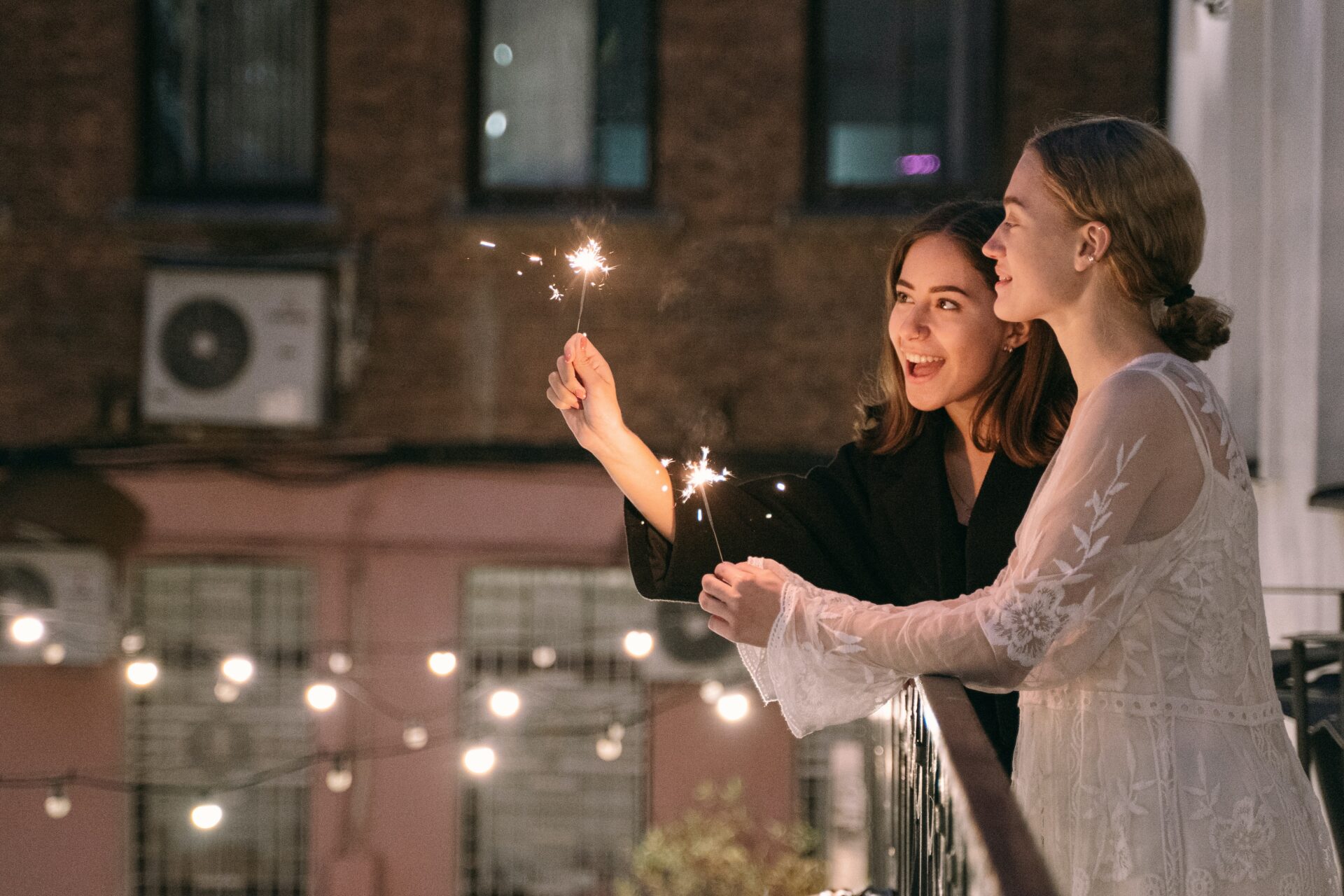 Two brides standing on balcony with cafe lights below