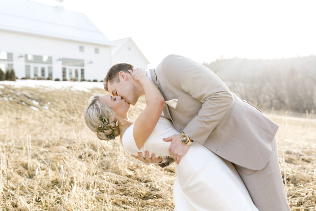 Fine art wedding photographer - groom dips and kisses bride in front of beautiful white barn