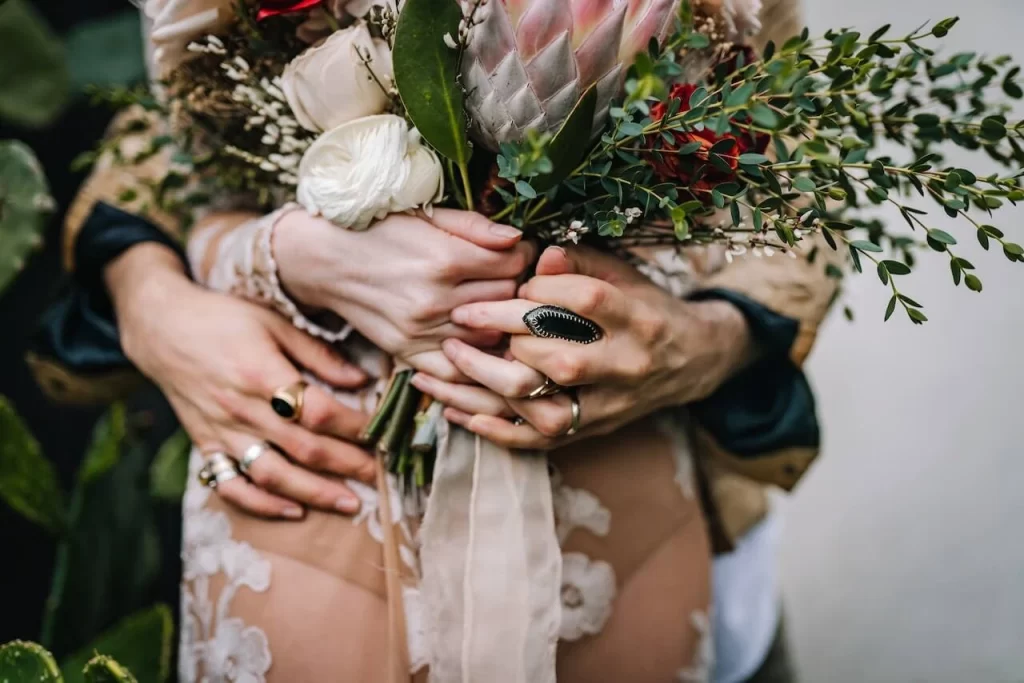 groom holding bride from behind with their hands grasping boho wedding bouquet for portrait to be featured on wedding directories